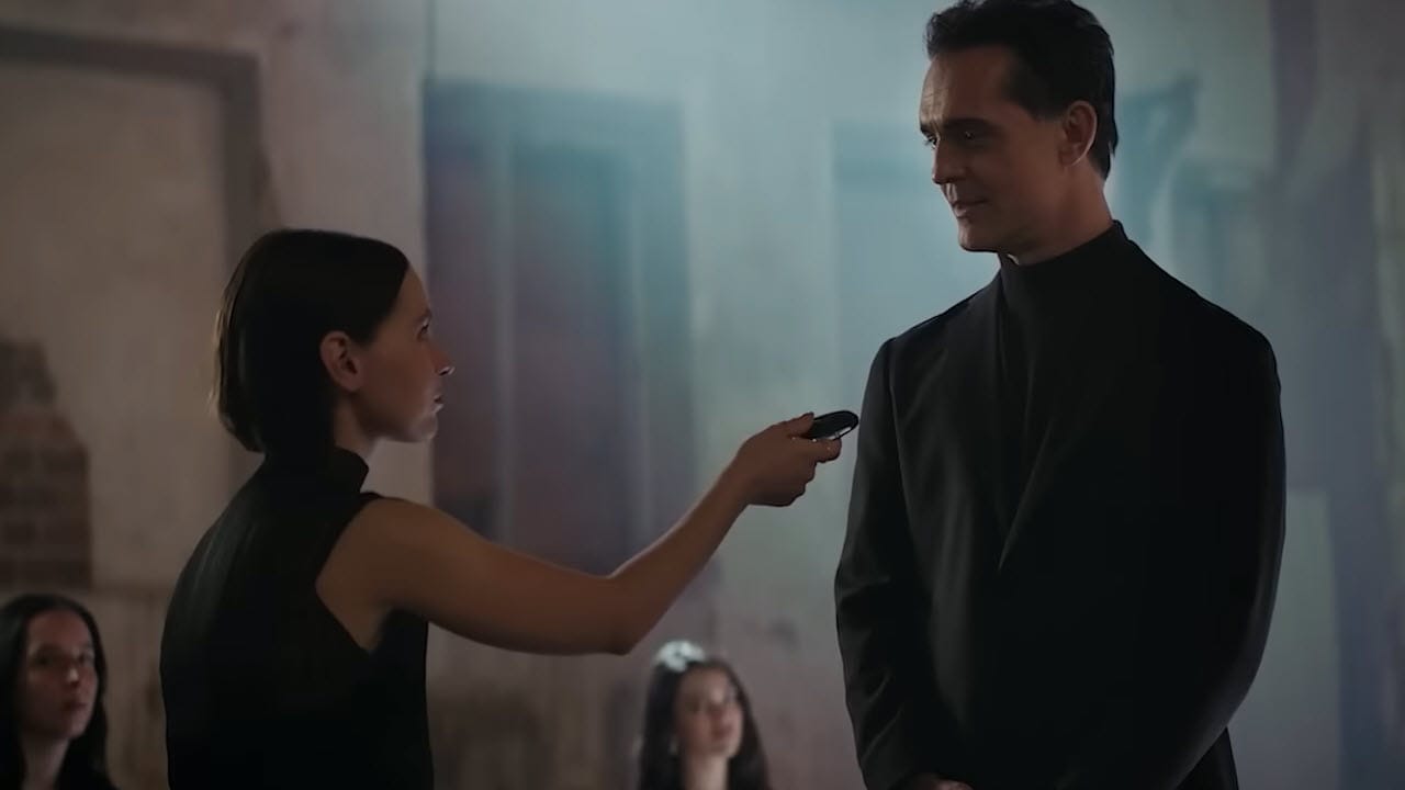 A woman in black holds a microphone toward a man in black standing beside her, with two seated women visible in the blurred background.