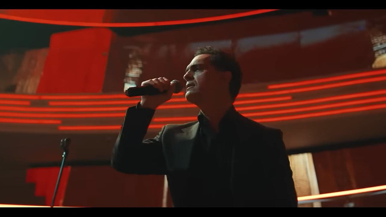 A man in a dark suit holds a microphone and sings on stage, illuminated by red curved lights in the background.