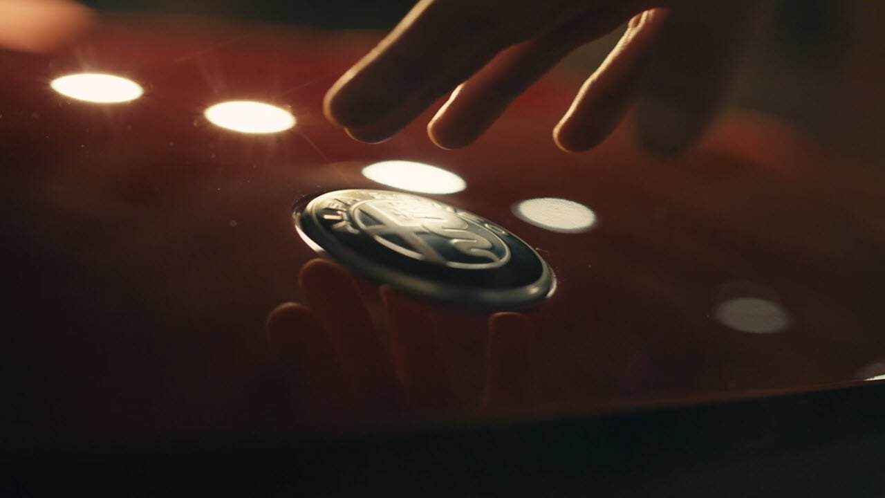 A hand reaches towards the Alfa Romeo logo on the hood of a red car, with round lights reflected on the surface.