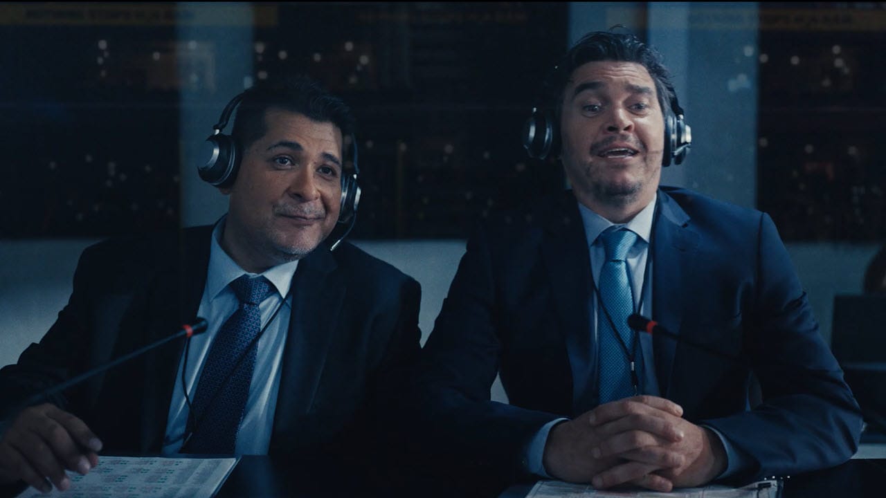Two men in suits wearing headsets sit at a desk with microphones and papers, appearing to commentate or report in a broadcast studio.