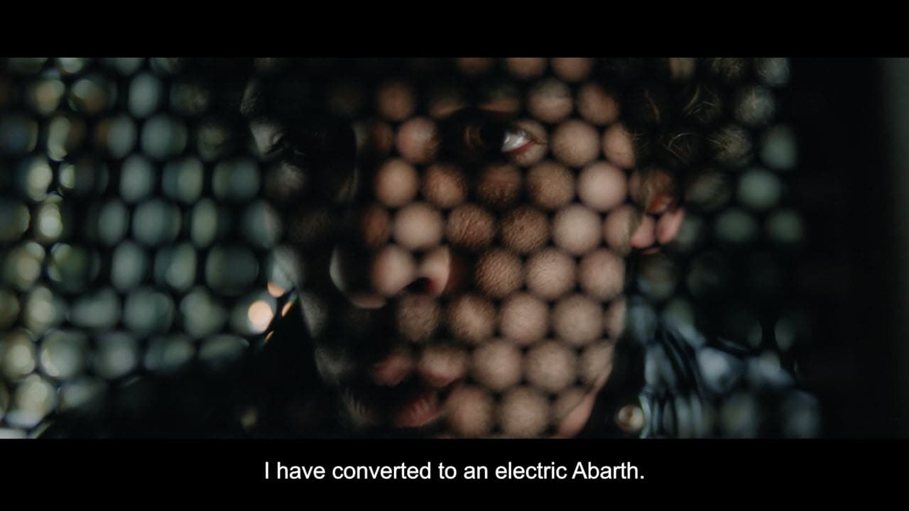 Close-up of a person’s face seen through a perforated screen, with the subtitle “I have converted to an electric Abarth.”.