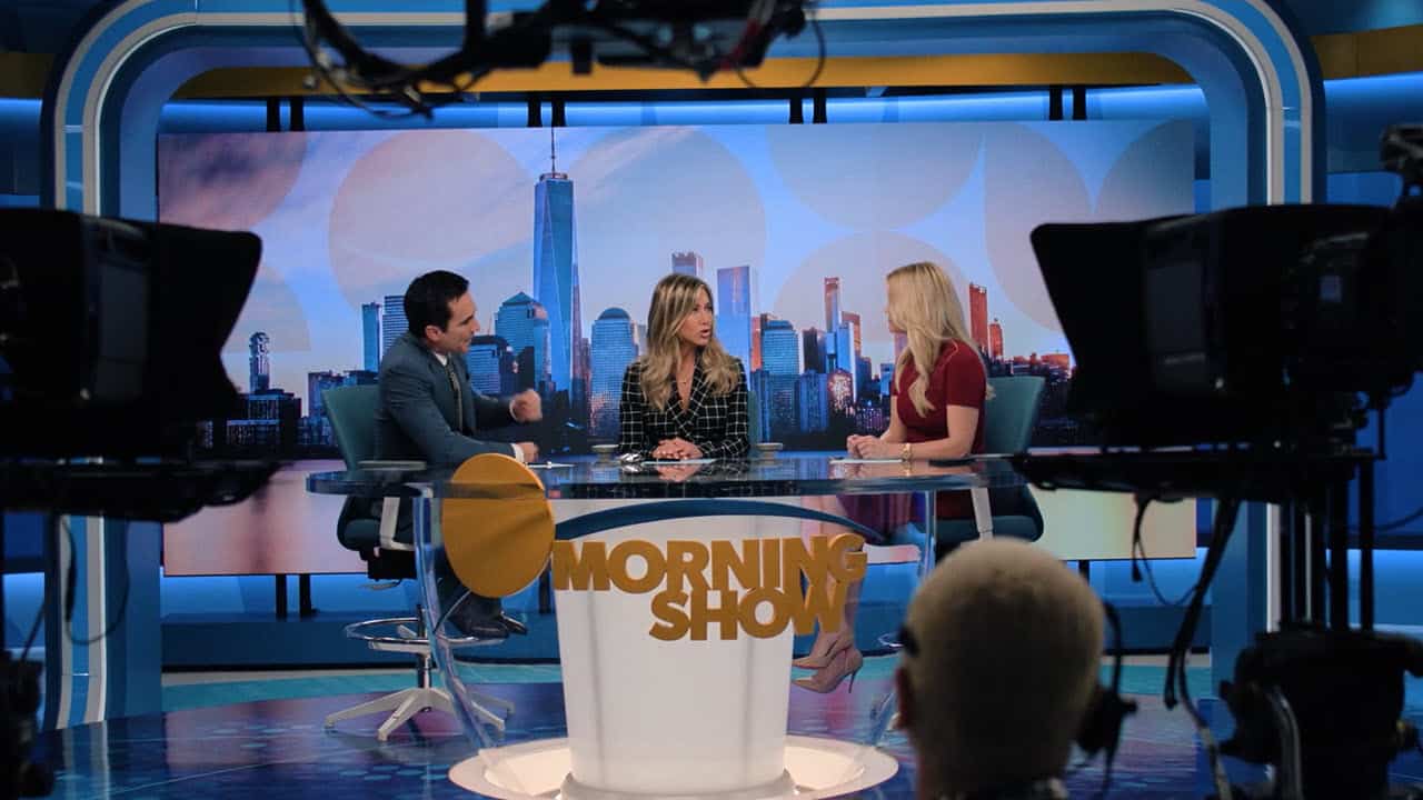 Three hosts sit at a desk on the set of a TV morning show, with cameras filming and a city skyline backdrop behind them.